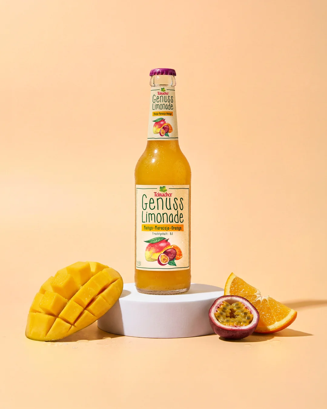 Flasche Teinacher Genuss Limonade Mango-Maracuja-Orange auf einem weißen Sockel, umgeben von einer aufgeschnittenen Mango, Orangenspalte und halbierter Maracuja vor pfirsichfarbenem Hintergrund