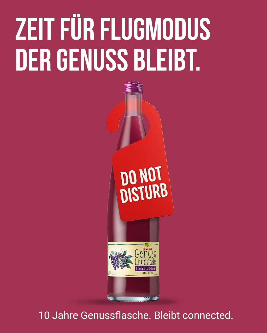 Teinacher-Kampagnenmotiv 'Zeit für Flugmodus – der Genuss bleibt': Flasche Teinacher Genuss Limonade Johannisbeer-Holunder mit rotem 'Do Not Disturb'-Türanhänger vor bordeauxrotem Hintergrund