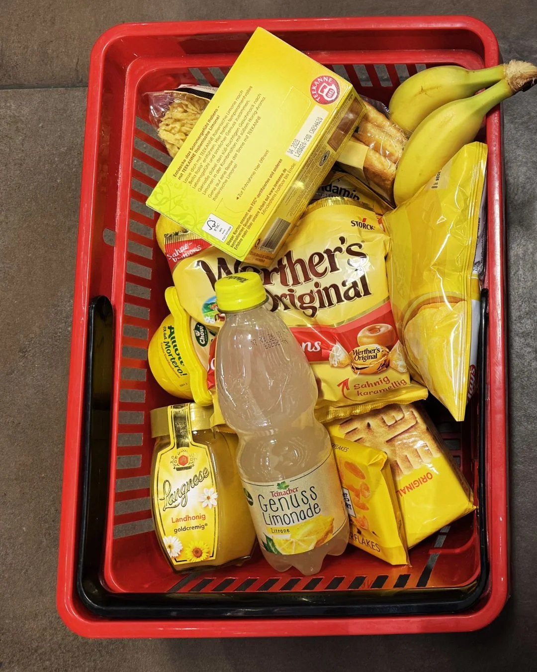 Einkaufskorb gefüllt mit ausschließlich gelb verpackten Produkten: Bananen, Honig, Cornflakes, Werther's-Original-Bonbons und eine Flasche Teinacher Genuss Limonade Zitrone