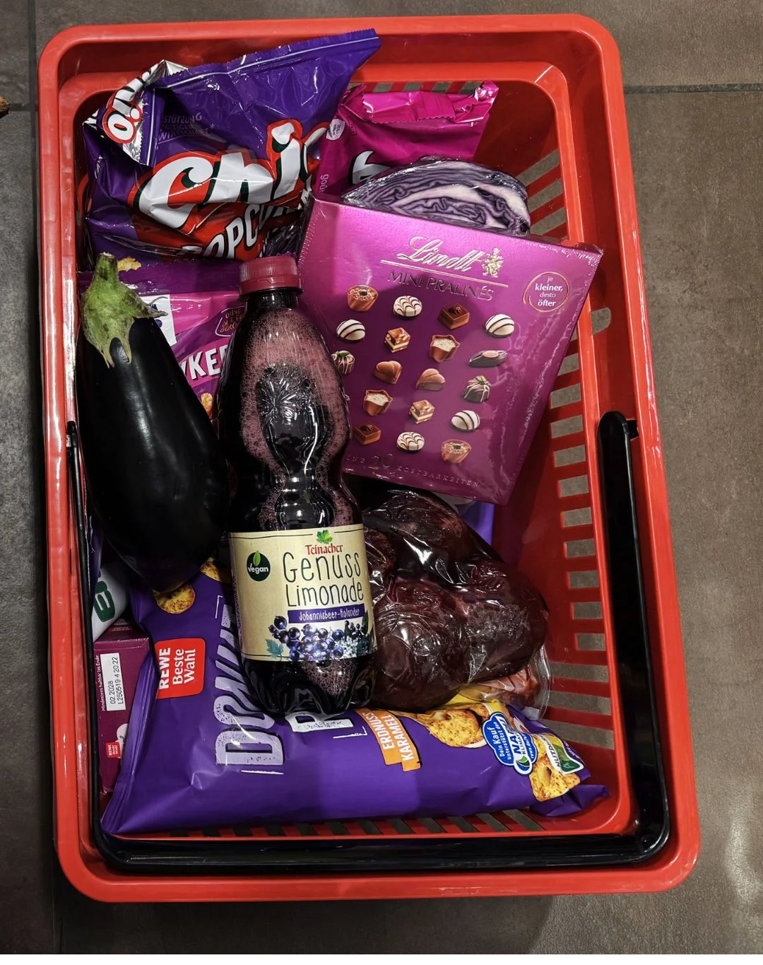 Einkaufskorb gefüllt mit ausschließlich violett verpackten bzw. violetten Produkten: Popcorn, Pralinen, eine Aubergine, Weintrauben, Kekse und eine Flasche Teinacher Genuss Limonade Johannisbeer-Holunder